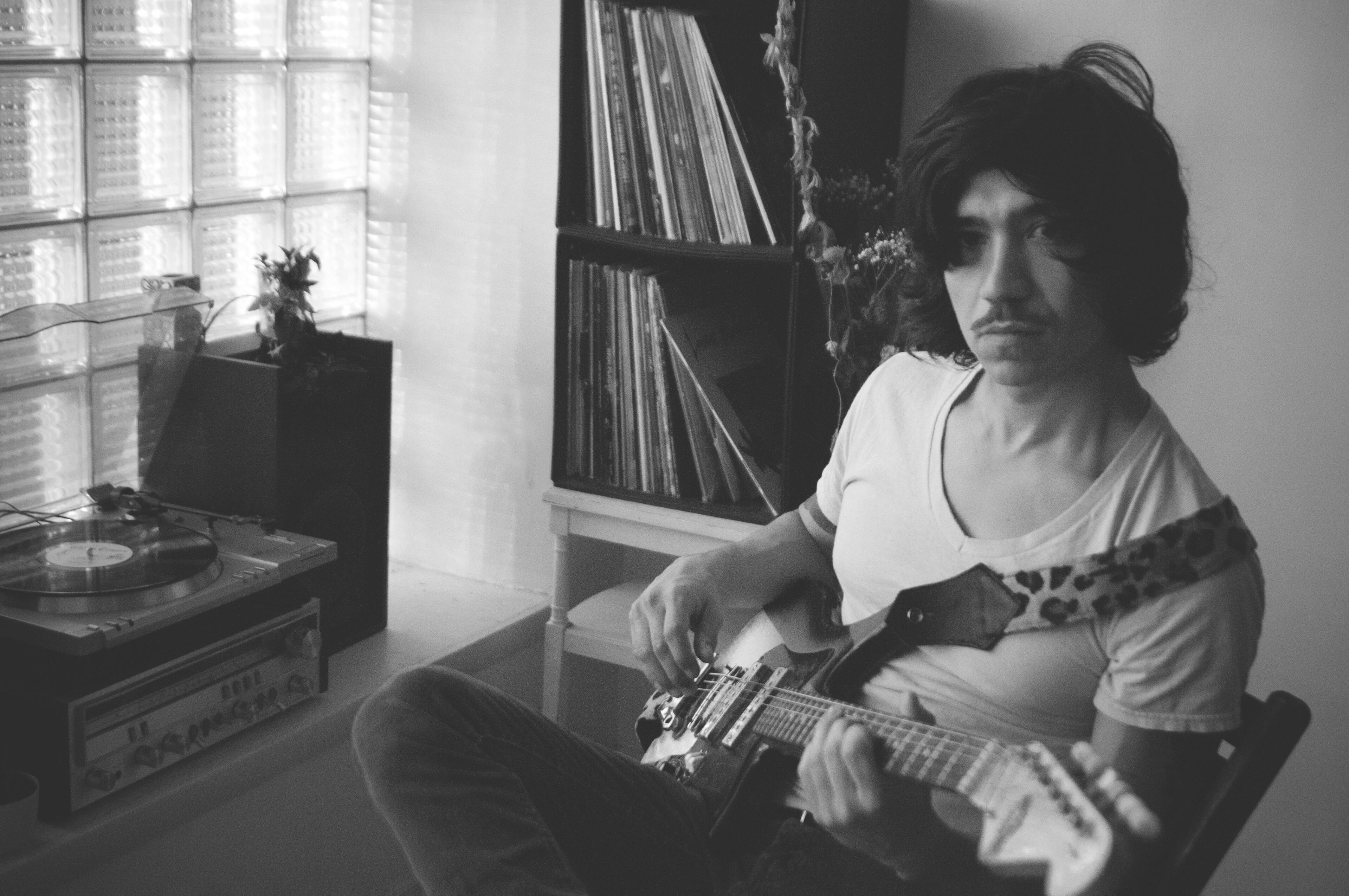 photo by Ashleigh Dye A black and white photo of Dan Rico sitting by a block-glass window with guitar, surrounded by records and a record player.